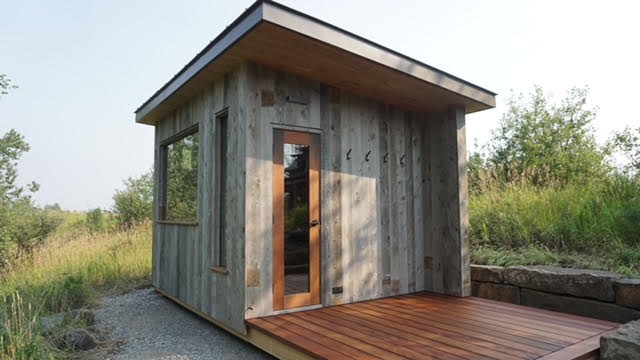 Weathered wood-sided sauna with a wraparound cedar deck, screen door, and gravel yard